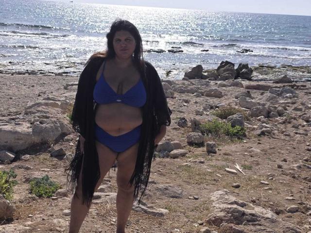 YasminQueen è in piedi su una spiaggia rocciosa, indossa un bikini blu con uno scialle nero frangiato, mettendo in mostra il suo seno grande e cadente e il suo corpo formoso e robusto, mentre la luce del sole si riflette sull'oceano..