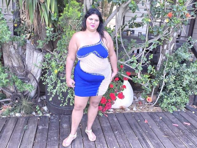 YasminQueen, con il suo corpo formoso e la pelle colorata, i capelli neri e ondulati, indossa un abito asimmetrico blu e beige, è in piedi su un ponte di legno, dietro di lei ci sono rose rosse in un vaso bianco..