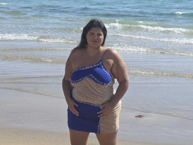 YasminQueen, con grandi seni, capelli scuri, indossa un abito scintillante blu e oro, in piedi su una spiaggia con le mani sui fianchi, l'oceano sullo sfondo..