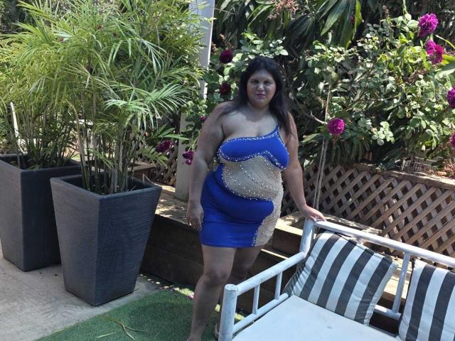 YasminQueen, con un sedere grande e un seno prosperoso, indossa un abito blu e beige, è in piedi in un giardino, appoggiata a una sedia a righe, ha i capelli scuri e sta sorridendo..