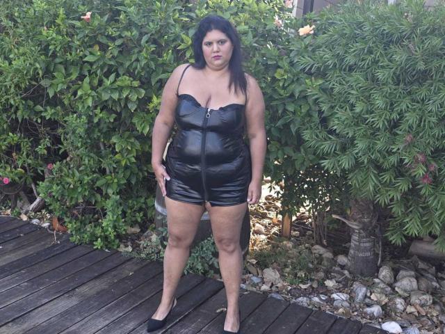 YasminQueen, eine übergewichtige Frau, steht im Garten, trägt einen schwarzen, glänzenden Overall mit Reißverschluss, schwarze High Heels, hat dunkles Haar, eine mittelgroße Brust, ist leicht mollig, im Hintergrund befindet sich ein grüner Busch und ein Holzboden..