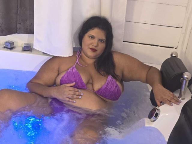 YasminQueen, plus-size woman with dark hair, in purple bikini, sitting in blue-lit bathtub, hand on large boobs, smiling, right arm on tub edge, white towel in background.