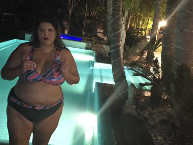 YasminQueen está em uma piscina à noite, vestindo um biquíni colorido, exibindo seus seios grandes e nádegas, com as mãos perto do peito, cabelos escuros e uma estrutura física ligeiramente acima do peso..