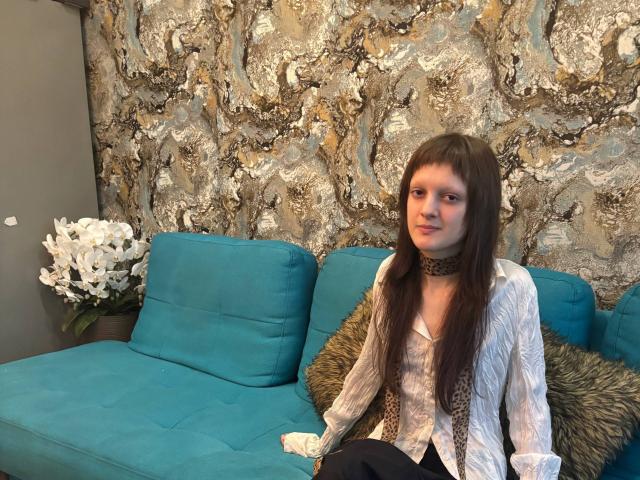 LisaGrenn with long brown hair, wearing a leopard print choker and white blouse, sits on a teal sofa with a furry pillow, looking slightly to the side.