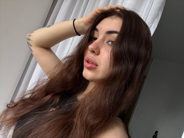 RaynaPeng with long brown hair, slightly messy, hand in hair, black sleeveless top, tattoo on arm, looking to side, pink lips, light skin, subtle makeup, white curtain in background.