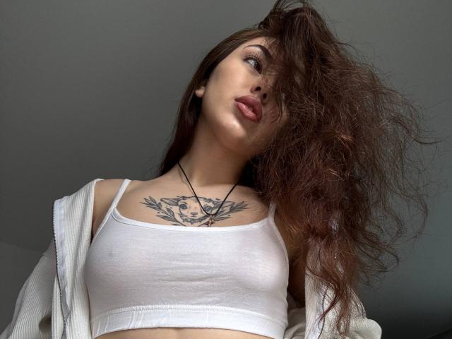 RaynaPeng with wavy brown hair, white tank top, and off-shoulder sweater, showing a tattoo on her chest, slightly parted lips, and a black necklace.