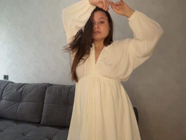 Evelina Newsom, con cabello castaño largo, vestida con un vestido blanco de manga larga y con botones, está de pie con los brazos levantados, mostrando sus senos y el contorno de su área genital, frente a un sofá gris..