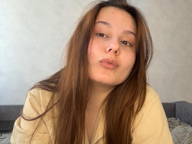 EvelinaNewsom with long brown hair, pouty lips, and a slightly tilted head, wearing a yellow shirt, looking directly at the camera.