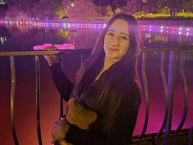 Omella, con el pelo largo y castaño, sonriendo, sosteniendo un cojín marrón y peludo, vistiendo una blusa negra, de pie junto a una barandilla de metal, con luces moradas y rosas en el fondo..
