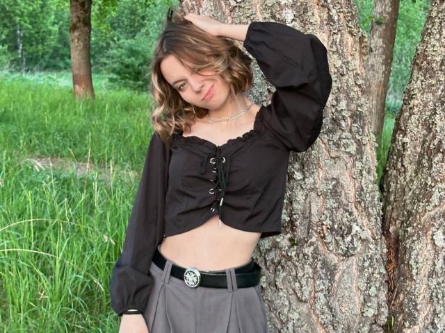 BrigetteLaurie with wavy brown hair, black off-shoulder blouse, and gray skirt, leaning against a tree, hand in hair, smiling, wearing a silver necklace and black belt.