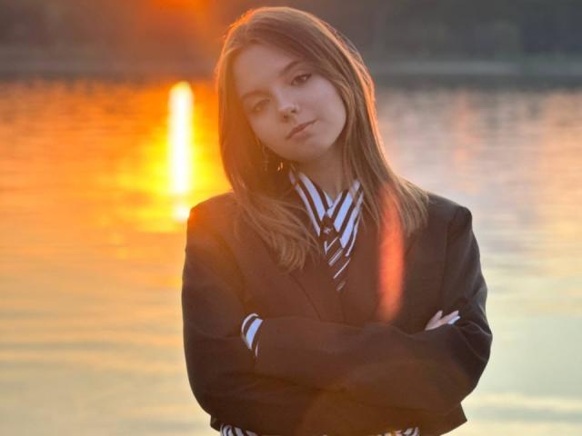 BrigetteLaurie with crossed arms, wearing a black suit jacket and striped shirt, standing by a sunset-lit water reflection, looking slightly annoyed. Her tits are small, and she's cute.