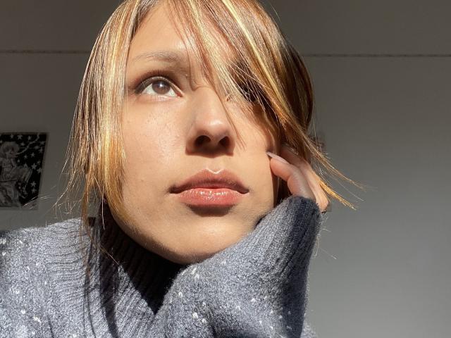 KiaraHernandezX with shiny blonde and brown hair, looking up, sunlight on her face, wearing a grey turtleneck, hand near her cheek, brown eyes, glossy pink lips.
