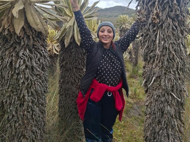 LizBonnet stands between tall palm trees with arms raised, wearing a gray beanie, black polka-dot shirt, and red jacket tied at the waist, smiling with dark eyes.