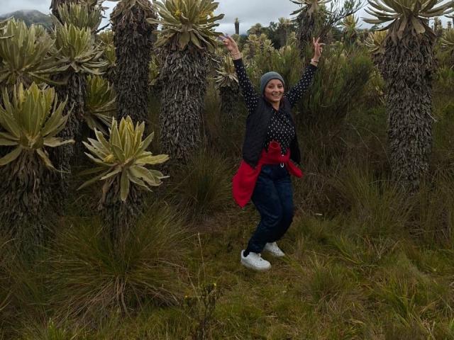 LizBonnet with arms up, smiling, wearing a blue headscarf, black polka dot shirt, red vest, blue jeans, and white shoes, standing among tall, spiky green plants.