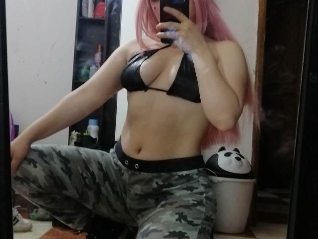 Nyxaria, com cabelo rosa, top de biquíni preto que mostra seus seios, e calças de camuflagem, agachada, tirando uma selfie no espelho, segurando um celular, em um quarto bagunçado, com uma máscara branca..