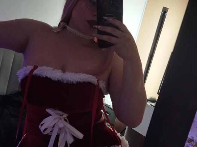 Nyxaria mit rosa Haaren, trägt ein rotes Samt-Kleid ohne Träger mit weißem Fellbesatz und einem weißen Band und macht ein Selfie im Spiegel, auf dem ihr Dekolleté und roter Lippenstift zu sehen sind..