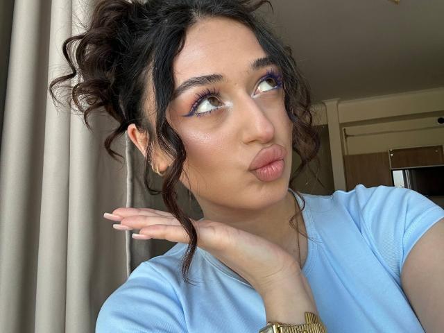 MyrtieGolds with curly dark hair in a messy bun, blue eyeshadow, pink lips, pouty expression, hand near face, wearing a light blue shirt, gold watch, looking to the side.
