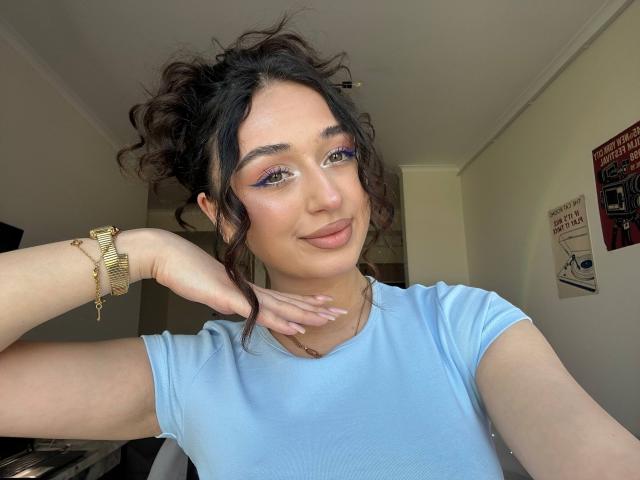 MyrtieGolds with curly brown hair, blue eye makeup, and pink lipstick, wearing a light blue shirt, gold watch, and necklace, smiling at the camera.