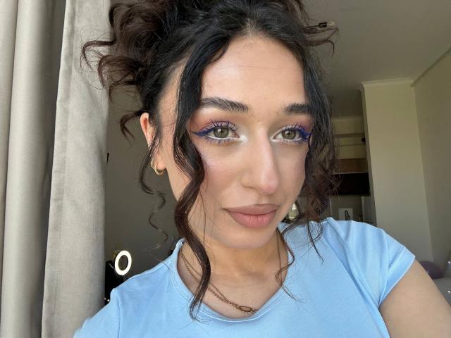 MyrtieGolds has wavy brown hair, blue eyeshadow, and a light blue shirt, with a slight smirk, looking at the camera. Her tits are small and her ass is unseen.