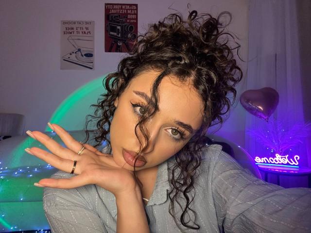 MyrtieGolds with curly brown hair, gray eyes, and pink lipstick, wearing a gray shirt, hand near face, neon lights in background, ring on finger, subtle purple and blue glow.