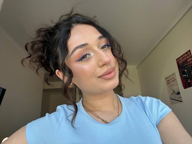 MyrtieGolds has curly dark hair, blue eyeshadow, and a light pink nose. She's wearing a light blue shirt, smiling slightly, with a subtle makeup look.