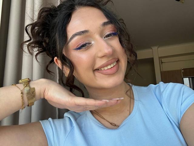 MyrtieGolds with curly dark hair, blue eyeshadow, and pink lipstick, smiling with tongue out, wearing a gold watch and light blue shirt, hand near face.