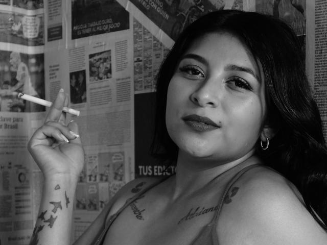Black-and-white photo of ArianaFoxxy with long dark hair, holding a cigarette, tattoos on her chest and arm, hoop earrings, smiling, wearing a tank top, looking at the camera.