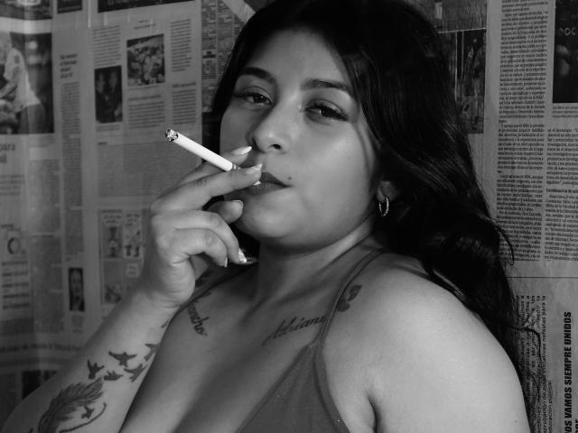 ArianaFoxxy with dark hair, smoking a cigarette, wearing a tank top, showing tattoos on her arm and chest, with hoop earrings, in a black-and-white photo.