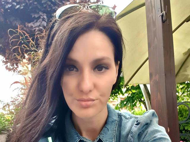 JoliDanielle tem cabelos escuros e ondulados, olhos castanhos e um sorriso discreto. Ela veste uma jaqueta jeans, com óculos de sol na cabeça, exibindo um visual adorável e jovial..