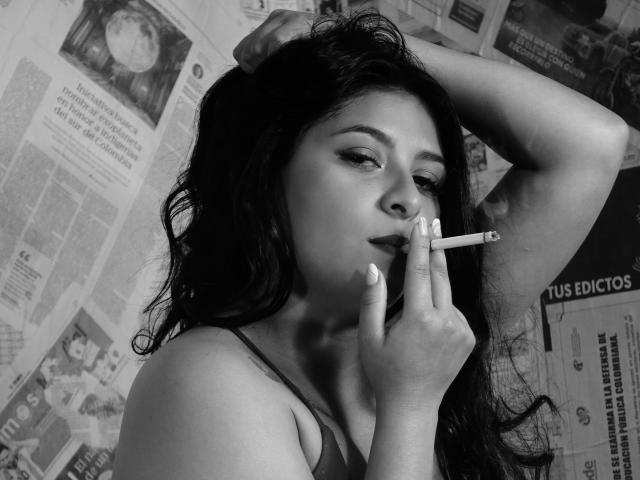 ArianaFoxxy with long dark hair, smoky eyes, and glossy lips, holding a cigarette to her slightly parted lips, her right hand raised behind her head. Black and white.