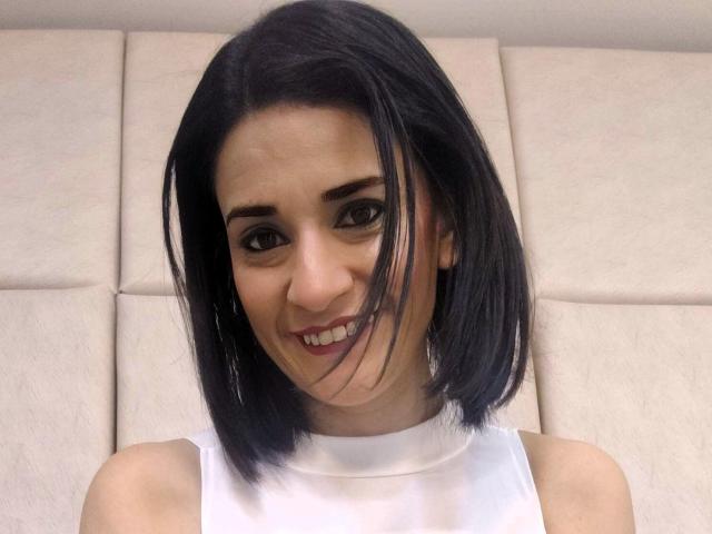 MaraRoberttss with short black hair, smiling, wearing a white sleeveless top, showing her tits slightly with her arms up, in a headshot.