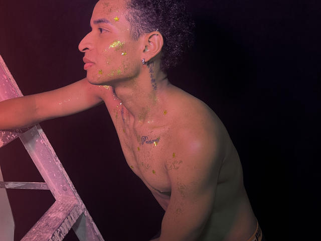 Chris, sin camisa, con manchas de pintura amarilla en la cara y el pecho, apoyado en una escalera, fondo oscuro, cabello corto y rizado, arete pequeño, tatuaje debajo de la barbilla, luz púrpura tenue..