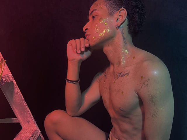 ChrisSexi com glitter no peito e no rosto, sem camisa, sentado com a perna levantada, mão no queixo, perto de uma escada, fundo escuro, usando uma pulseira preta, olhando de perfil..