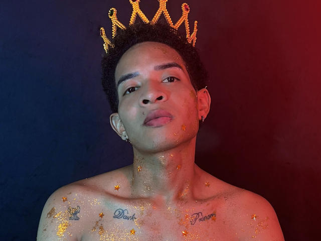 ChrisSexi met een gouden kroon, zonder shirt, oranje glitter op borst en gezicht, tatoeages op de schouders, kort krullend haar, rode en blauwe verlichting, kleine oorbellen, kijkt omhoog..