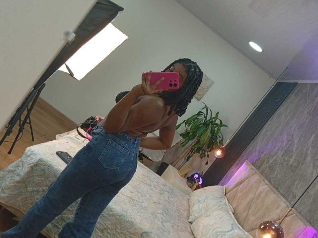 AmberrQueen with dark skin, braided hair, topless, showing her back, wearing blue jeans, holding a pink phone, reflecting in a mirror, bedroom, soft lighting, potted plant.