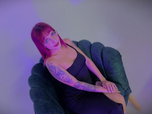 SachaNovak, con cabello rosa, ojos azules y tatuajes, vestida con un vestido negro, sentada en una silla de terciopelo verde, con las manos sobre la rodilla, iluminación morada y azul..