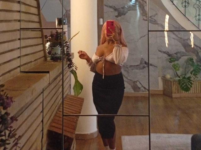 AmberrQueen has pink hair, a tied-up white top showing her boobs, and a tight black skirt. She's taking a mirror selfie with a red phone, holding a leaf.