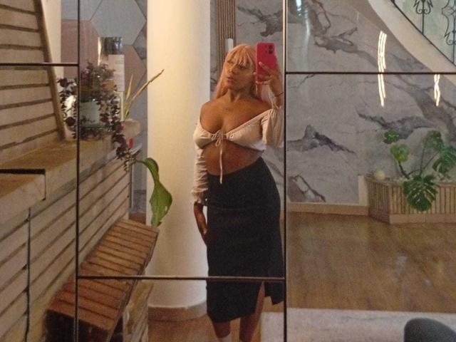 Mirror selfie of AmberrQueen with blonde hair, off-shoulder white top showing big boobs, black skirt with a thigh slit, holding pink phone, standing in wooden room.
