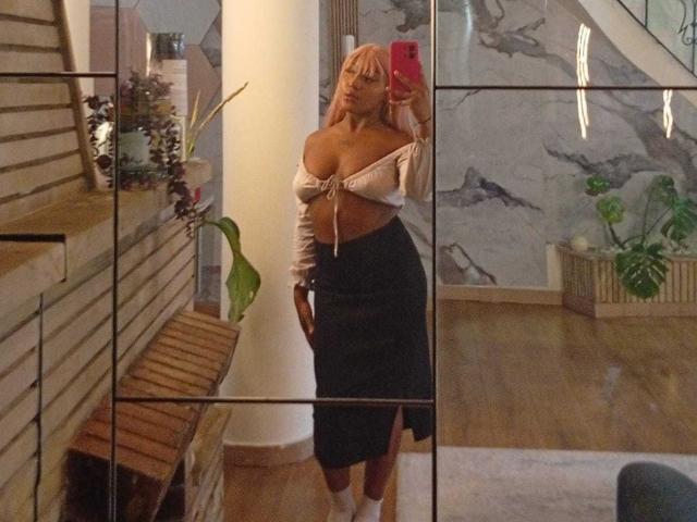 AmberrQueen with pink hair, off-shoulder white top tied below her tits, black skirt, standing in front of mirror, holding red phone, wooden floor, white column in background.