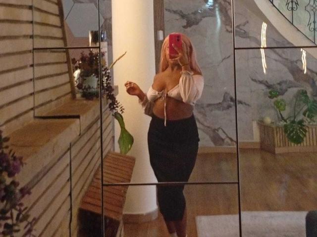 AmberrQueen with pink hair, large tits, tied-up white top, black skirt, standing in front of mirror, holding phone, reflecting in glass, wooden stairs, plant on side.