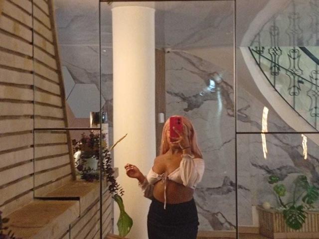 AmberrQueen with pink hair, off-shoulder top exposing her tits, holding a leaf, standing in front of a glass wall, wearing a black skirt, selfie in a modern room.