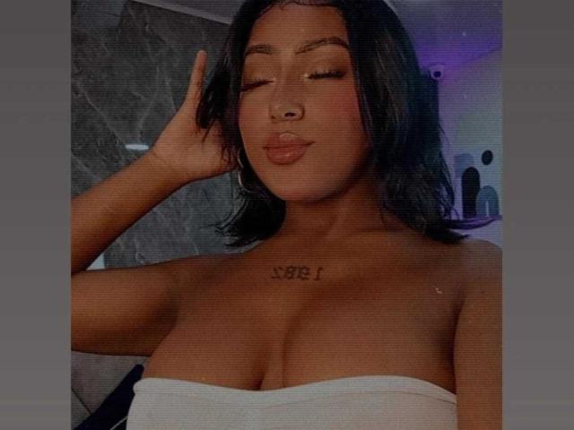 AmberrQueen with black shoulder-length hair, eyes closed, pouty lips, white strapless top, large boobs, chest tattoo "88" in bold, hand touching head, wearing hoop earrings.