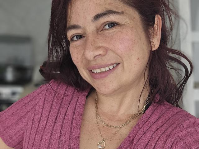 AlessiaRed with freckled face, smiling, wearing a pink ribbed shirt and gold necklace with a heart pendant, has medium brown hair, and blue eyes.