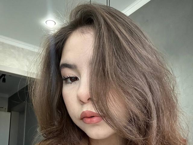 EmeraldCrivaro has wavy brown hair covering one eye, full red lips, and thick black eyelashes. Her pale skin and subtle makeup are visible. Her expression is neutral.