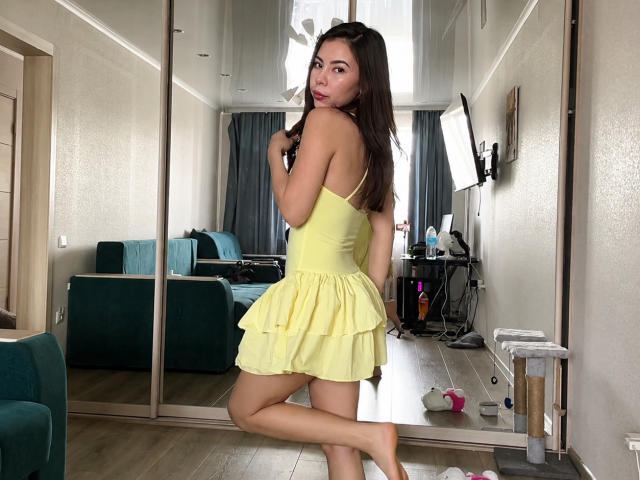 EmeraldCrivaro stands in a room, wearing a yellow dress, showing her ass, with her back to the camera, looking over her shoulder, dark hair, fair skin, wooden floor.
