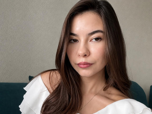 EmeraldCrivaro has smooth brown skin, long brown hair, brown eyes, and pink lipstick. She's wearing a white off-shoulder top, looking straight at the camera with a slight smile.