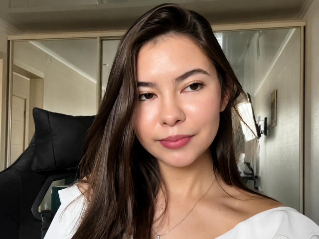 EmeraldCrivaro with long brown hair, light skin, and pink lips, wearing a white off-shoulder top, sitting in a black chair, looking at the camera with a slight smile.
