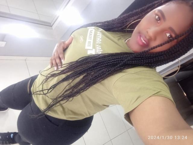 SkylerNaomi, avec de longues tresses, un t-shirt vert, un pantalon noir et des boucles d'oreilles cerceaux, la main sur ses grandes seins, souriante, debout dans une pièce carrelée, horodatée le 30/01/2024 à 13:24.