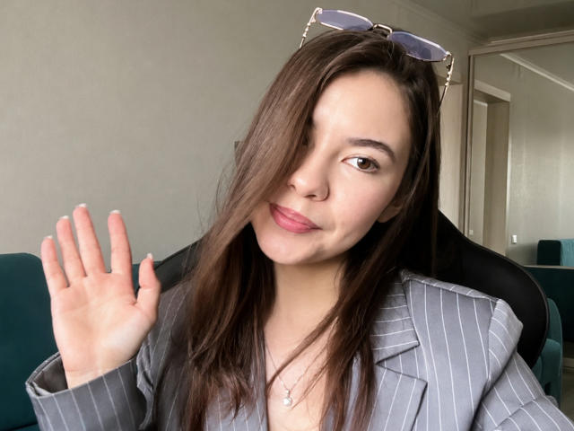 EmeraldCrivaro with long brown hair, wearing a gray pinstripe blazer, waving with her right hand, purple eyeglasses on her head, and a subtle smile, pink lips.