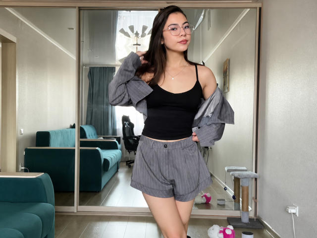 EmeraldCrivaro stands in a room, wearing a black tank top and gray pinstriped shorts, with a gray jacket off her shoulders, looking at the camera with glasses.
